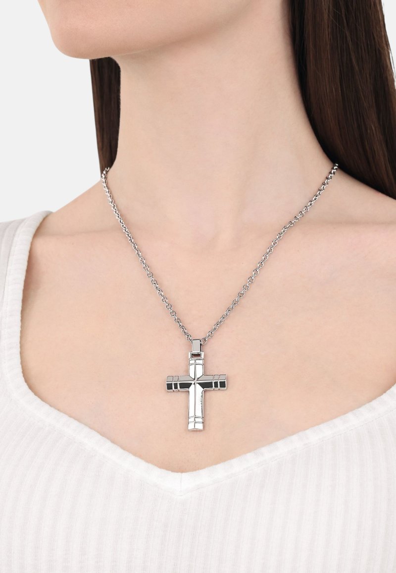 Silver crosshalsband med geometrisk design, med polerade och matta ytor, hängande på en länkad kedja, bärs över en vit ribbad topp.