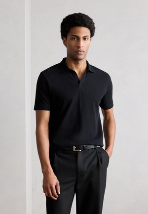 Egy rövid göndör hajú férfi, fekete rövid ujjú galléros pólót és fekete nadrágot visel, egyik kezét a zsebében tartva áll.