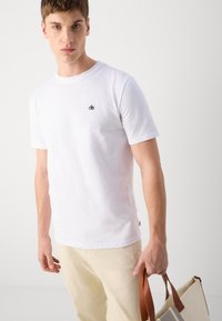 Hvid bomulds T-shirt med korteærmer, prydet med et lille sort logo på brystet. Båret med lyse bukser og holder en taske.