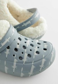 Lichtblauwe klompen met een zachte, witte fleece voering en een herhaald vospatroon. Bevat ventilatiegaten en een stevige constructie.