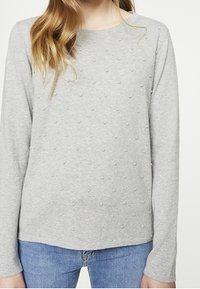 Nainen, joka pukeutuu vaaleanharmaaseen pitkähihaiseen neuleeseen, jossa on pieniä teksturoituja pisteitä, yhdistettynä sinisiin farkkuihin. Kuva on rajattu kaulasta lantioon.