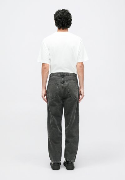 Mand med krøllet hår iført en hvid t-shirt, løse sorte jeans og sorte sko, stående vendt væk mod en ensfarvet hvid baggrund.