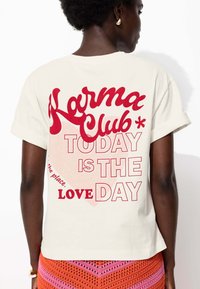 T-shirt en coton couleur crème avec un texte rouge : "Karma Club" et "AUJOURD'HUI EST LE JOUR DE L'AMOUR" au dos, avec des manches retroussées.