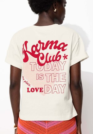 T-shirt en coton couleur crème avec un texte rouge : "Karma Club" et "AUJOURD'HUI EST LE JOUR DE L'AMOUR" au dos, avec des manches retroussées.