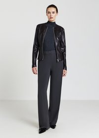 Veste en cuir noir de type bomber avec un col rond, portée avec un pull à côtes foncé et un pantalon gris ample. Chaussures noires brillantes.