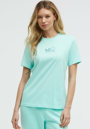 Blonde woman wearing a light turquoise t-shirt with "hi!" and smiley face text, paired with matching pants, standing against a plain background.