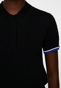 Svart pikétröja med ett strukturerat mönster, med en knappslå och korta ärmar med blå och vita randiga detaljer.