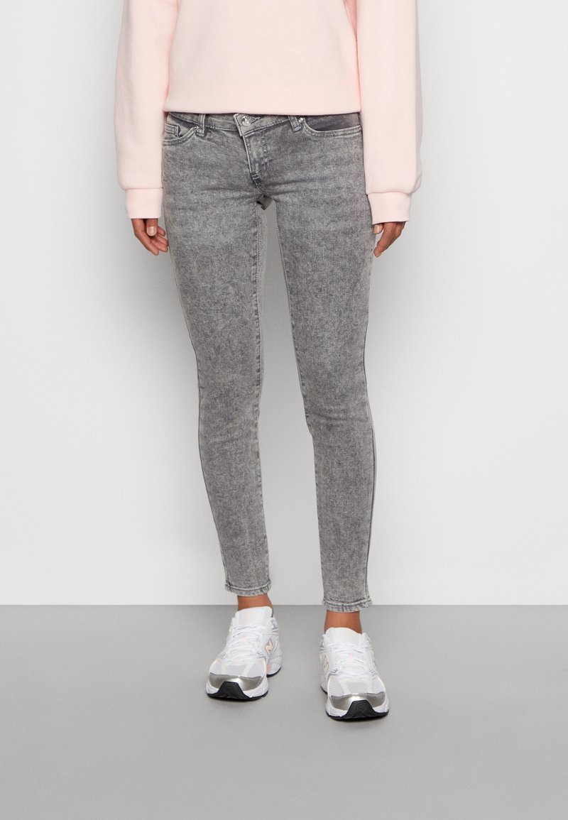 Jeans skinny grigi con un lavaggio sbiadito, vita media e dettagli di cucitura sottili, abbinati a scarpe da ginnastica bianche.