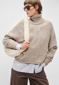 Beige pletený svetr s rolákem a texturovaným povrchem. V kombinaci se světle modrou košilí a hnědými kalhotami, drží krémovou kabelku z umělé kožešiny.