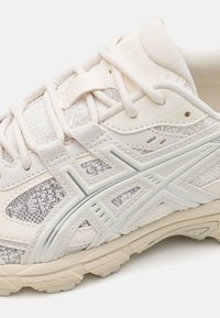 Beige athletic shoe with textured mesh and synthetic overlays, featuring a silver accent line and decorative stitching on the side.