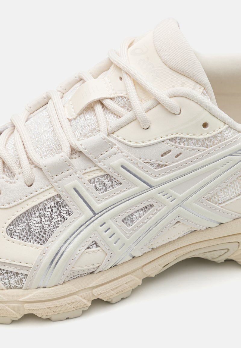 Beige Sport Schuh mit strukturiertem Mesh und synthetischen Überzügen, versehen mit einer silbernen Akzentlinie und dekorativen Nähten an der Seite.