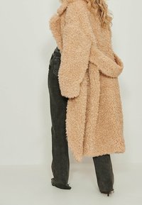 Nainen, joka käyttää mustia korkeavyötäröisiä farkkuja ja pitkää, beigeä, teksturoitua teddy-takkia, seisoo tasaisen vaalean taustan edessä.