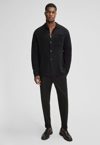 Czarna koszula na guziki z kieszonką, zestawiona z czarnymi spodniami i mokasynami; gładka faktura i luźny krój, podkreślający monochromatyczny wygląd.