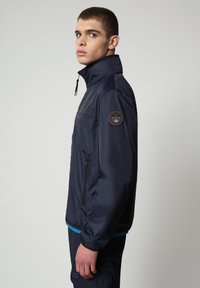 Giacca impermeabile navy con colletto alto, tasca laterale con zip e patch rotondo con logo sulla spalla sinistra. Texture liscia, design leggero.