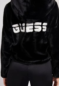 Black faux fur hoodie with a prominent white "GUESS" logo on the back. The design features a hood and soft texture.
