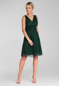 Grünes ärmelloses Kleid mit V-Ausschnitt, geraffter Taille und transparenter Überlage. Die A-Linien-Silhouette reicht bis zum Knie. Getragen mit klaren High Heels.