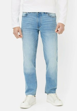 Mężczyzna w jasnoniebieskich dżinsach, białej koszuli z długim rękawem i białych trampkach, stojący z rękami swobodnie opuszczonymi wzdłuż ciała.