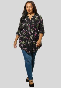 Tunic nera a fiori con bottoni frontali, maniche a 3/4 e dettagli plissettati; abbinata a jeans skinny blu e scarpe flat nere.