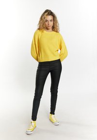 Gelbes, geripptes Strickpullover, schwarze eng anliegende Hose und gelbe High-Top-Sneaker mit weißen Sohlen. Lockige Haare, Arme entspannt an den Seiten.