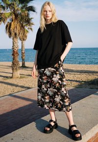 Blond kvinde står nær strandpalmer iført sort oversized t-shirt, floral midi nederdel og sorte chunky sandaler under klar himmel.