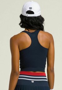 Top corto de color navy con textura en relieve y diseño de espalda nadadora; combinado con una falda con pliegues que presenta rayas rojas y blancas en la cintura. Gorra blanca con logo.