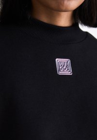 Čierna mikina s rebrovaným golierom, so strieborným a ružovým kovovým štvorcovým logom na hrudi. Hladká textura látky.
