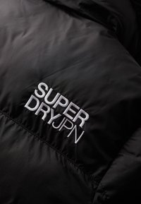 Zwarte stof met witte geborduurde tekst "SUPER DRY JPN" op een doorgestikte oppervlakte, met een accent op de details van het kledingmerk.