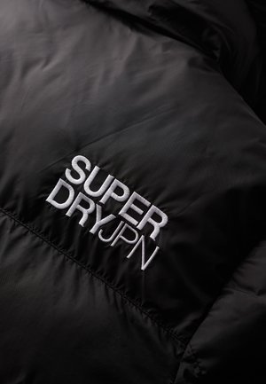 Czarna tkanina z białym haftem "SUPER DRY JPN" na pikowanej powierzchni, podkreślająca szczegóły marki odzieżowej.