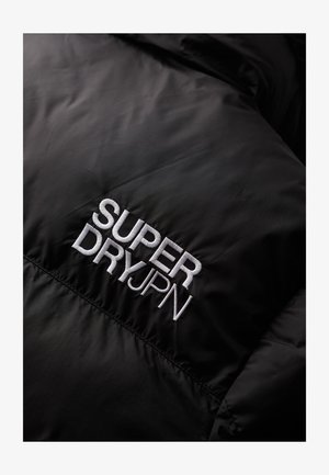 Schwarzer Stoff mit weißem gesticktem Text "SUPER DRY JPN" auf einer gesteppten Oberfläche, der das Detail der Bekleidungsmarke hervorhebt.