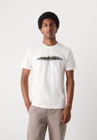Hvit bomulls t-skjorte med en trykket logo i svart og grønt, med dryppende malereffekt, sammen med lyse beige bukser.