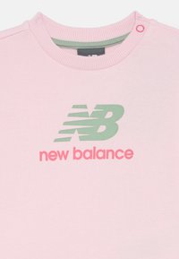 Lichtroze New Balance sweater met ribbelboord aan de hals, groen en roze logo gedrukt op de borst, en een drukknoop op de schouder.
