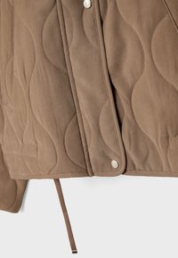 Časť hnedej prešívanej bundy zobrazujúca patentky, vlnovité prešívanie a visiaci textilný sťahovací šnúrku.