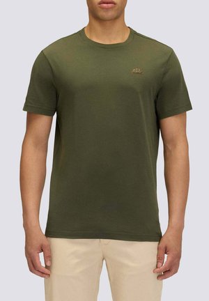 Uomo con t-shirt verde oliva a maniche corte e pantaloni beige chiaro, in piedi davanti a uno sfondo grigio chiaro uniforme.