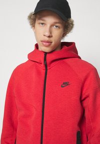 Felpa rossa Nike con chiusura a zip, dettagli neri e logo. Presenta un cappuccio arrotondato e un tessuto testurizzato. Inclusi taschini laterali con zip.