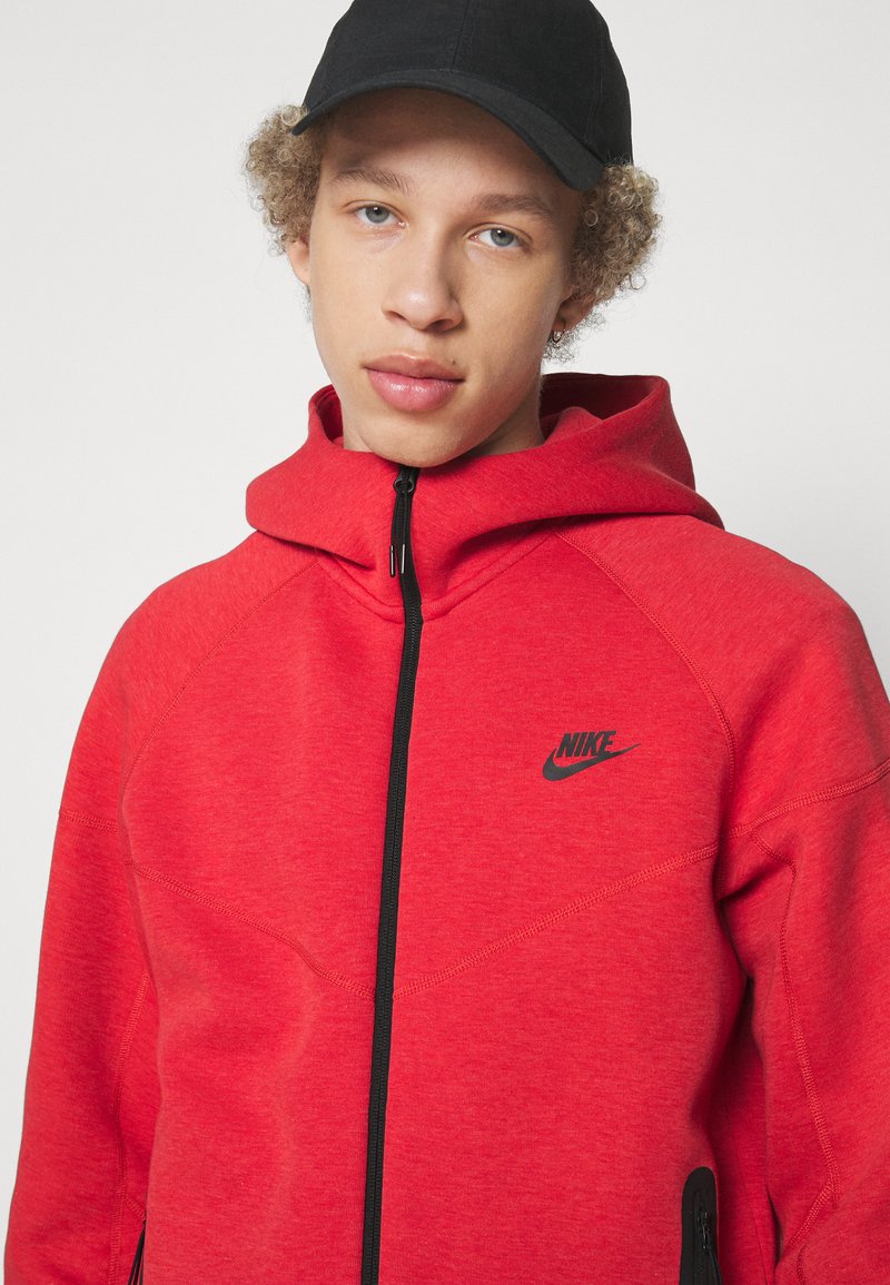 Czerwona bluza Nike z kapturem na zamek, z czarnymi akcentami i logo. Posiada zaokrąglony kaptur i teksturowany materiał. Zawiera boczne kieszenie na zamek.