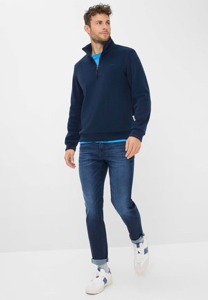 Marineblauer Fleecepullover mit hohem Kragen, Reißverschluss, gerippten Bündchen und Saum; getragen über einem hellblauen T-Shirt mit dunkelblauer Jeans und weißen Sneakers.