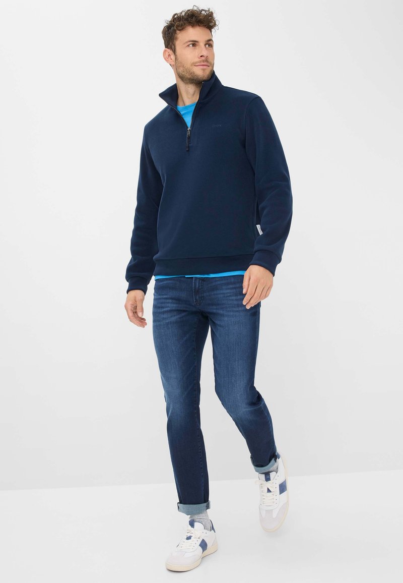 Maglione in pile blu navy con collo alto, chiusura con zip, polsini e orlo a coste; indossato sopra una t-shirt azzurro chiaro con jeans scuri e scarpe da ginnastica bianche.