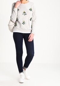 Sudadera gris con patrones de árboles verdes y amarillos, cuello redondo y puños acanalados, combinada con jeans ajustados oscuros y zapatillas blancas.