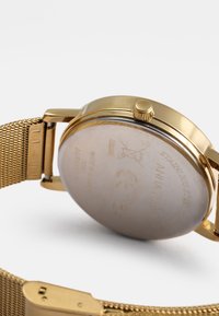 Χρυσό ρολόι από ανοξείδωτο ατσάλι με πλέγματα. Στρογγυλή θήκη και υφασμένη κορώνα. Ο εγχαραγμένος πίσω δείχνει λεπτομέρειες της μάρκας και της κατασκευής.