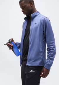 Αθλητικός άνδρας που φορά ένα μωβ ζακέτα με φερμουάρ και μαύρο παντελόνι Nike Pro, κρατώντας ένα μπλε και μαύρο παγούρι νερού, κοιτώντας προς τα κάτω.