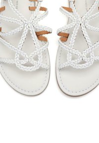 Sandalias trenzadas blancas con acentos de cuero, suela plana, diseño de tiras intrincado y detalles de costura visibles sobre un fondo neutral.