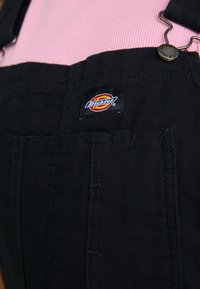 Gros plan sur une salopette noire Dickies portée par-dessus un t-shirt côtelé rose, montrant l'étiquette du logo et la boucle métallique sur la bretelle.