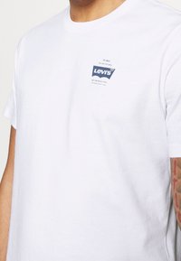 Bílá bavlněná tričko s modrým logem Levi's na levé části hrudníku, kulatým výstřihem a krátkými rukávy. Měkká, hladká textura.