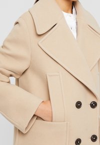 Abrigo de mezcla de lana beige con diseño de doble botonadura, grandes solapas, bolsillos frontales y botones oscuros, con un acabado suave y texturizado.