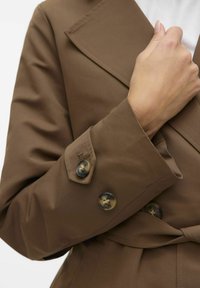 Brun trenchcoat med en bred krage. Funktioner två mörka knappar på ärmarna, texturerat tyg och ett knytbart midjebälte för justering.