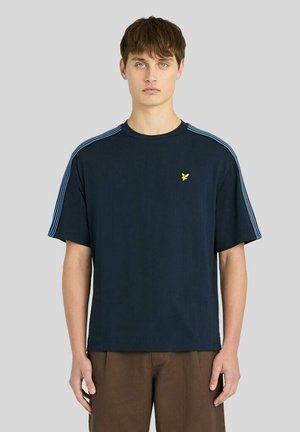Junger Mann trägt ein marineblaues T-Shirt mit hellblauen Streifen auf den Schultern und einem kleinen gelben Vogel-Logo auf der linken Brust.