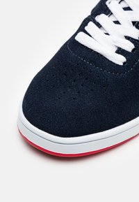 Mörkblå mockaskor med vita snörningar, rundad tå och rosa gummisula. Har perforeringar vid tån för ventilation.