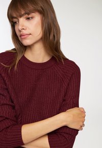 Burgundowy sweter z dzianiny o teksturowanym wzorze, z prążkowanymi mankietami i okrągłym dekoltem. Wykonany z miękkiego materiału, z rękawami raglanowymi i bezszwowym wykończeniem.