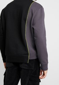 Sudadera en negro y gris oscuro, con un hilo de acento amarillo a lo largo de la costura. Hecha de una mezcla de algodón texturizado y con un corte relajado.