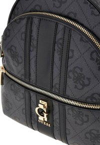 Zaino in tessuto nero con design del logo a fantasia, hardware dorato, chiusura zip e strisce texture. Presenta lettere in rilievo e dettagli. 
